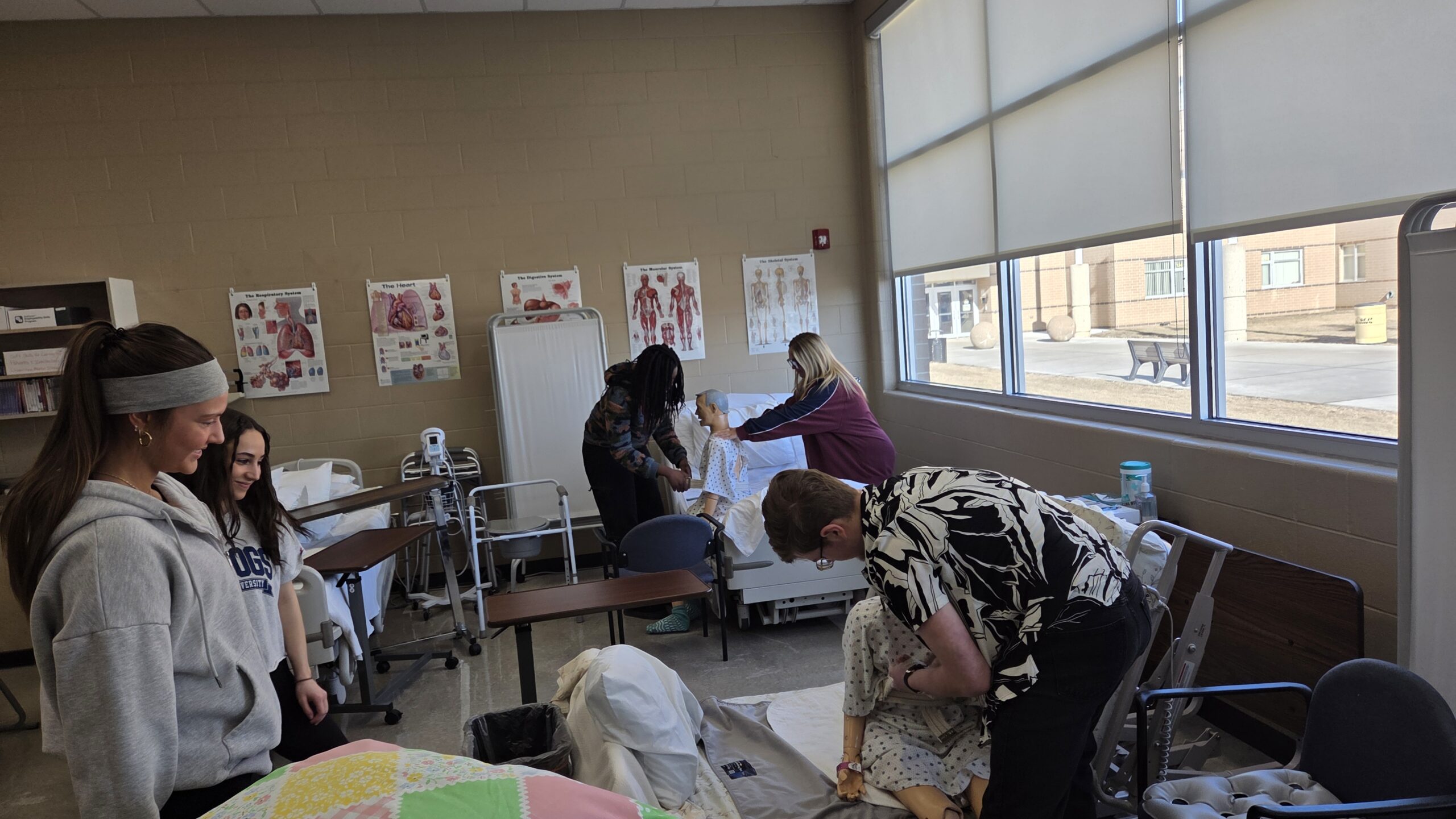 Students applying gait belts to the mannequins practicing transferring a resident from the bed to a chair. Students in the picture going clockwise, starting with the female wearing a gray headband, are Addilyn Larsen, Izabella Miller, Margaret Danso, Lily White, and Connor Warner.