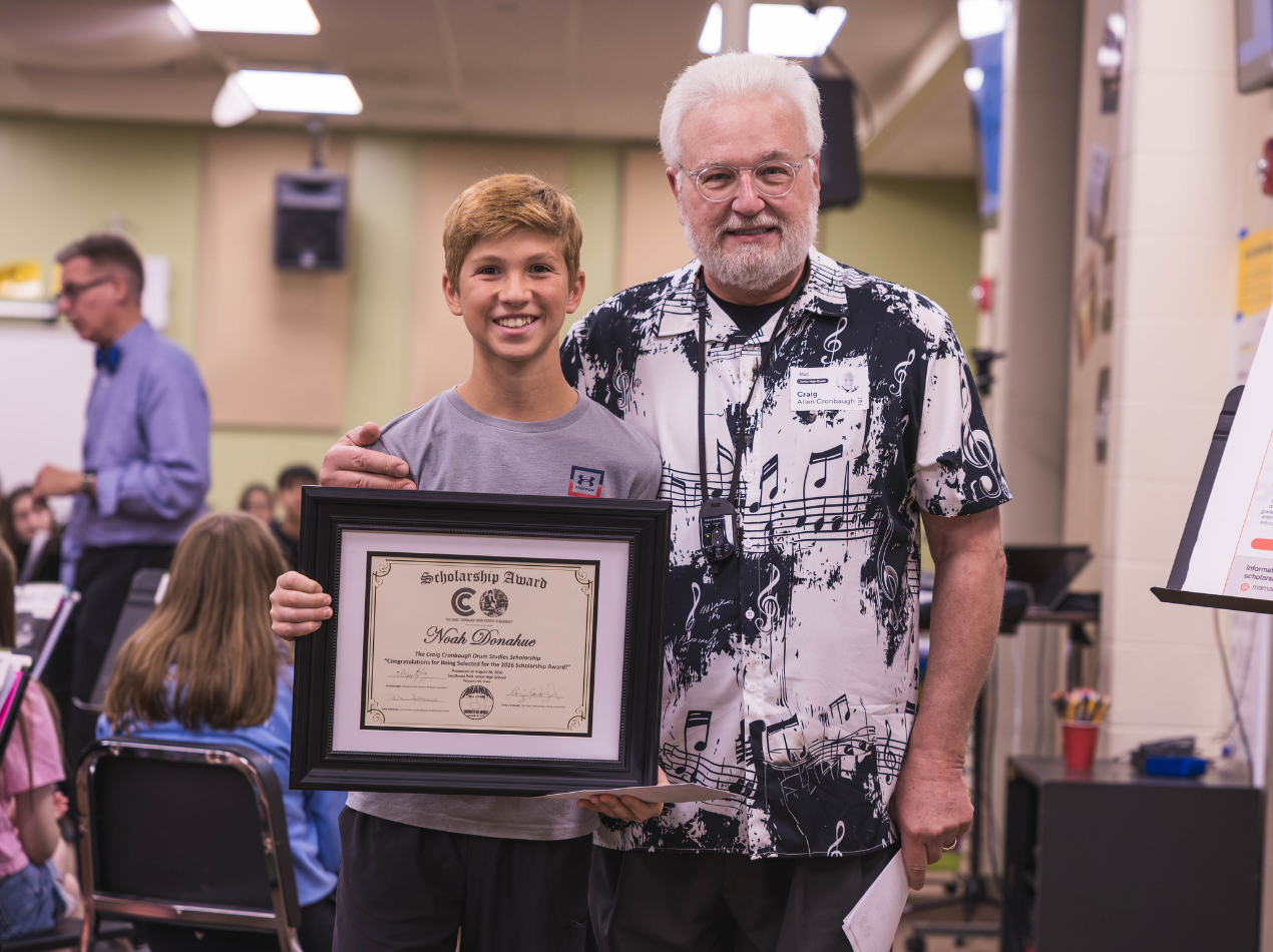 Two people stand in an indoor awards event; one holds a framed certificate while smiling, the other stands beside them with a white beard, both facing the camera under bright ceiling lights.