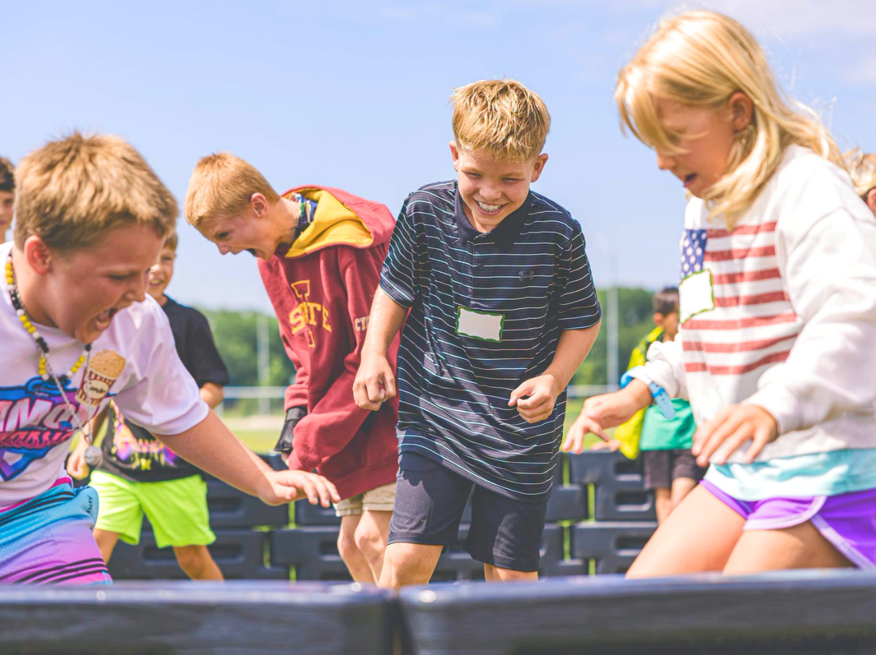 Children outdoors playing a running game on a track, smiling and reaching forward as they move past colorful blocks under a blue sky.
