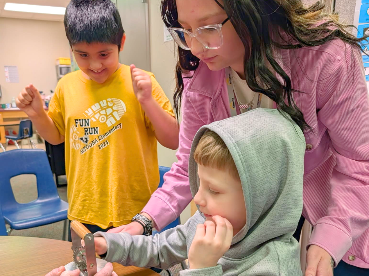 Adult and two children at a classroom table, the adult helping as a child in a green hoodie eats or tastes food while another child in a yellow shirt stands nearby.