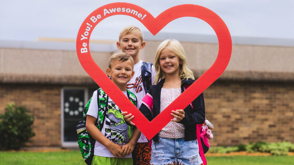 Three kids pose outdoors in front of a building, holding a large red heart shape with the text “Be You! Be Awesome!”