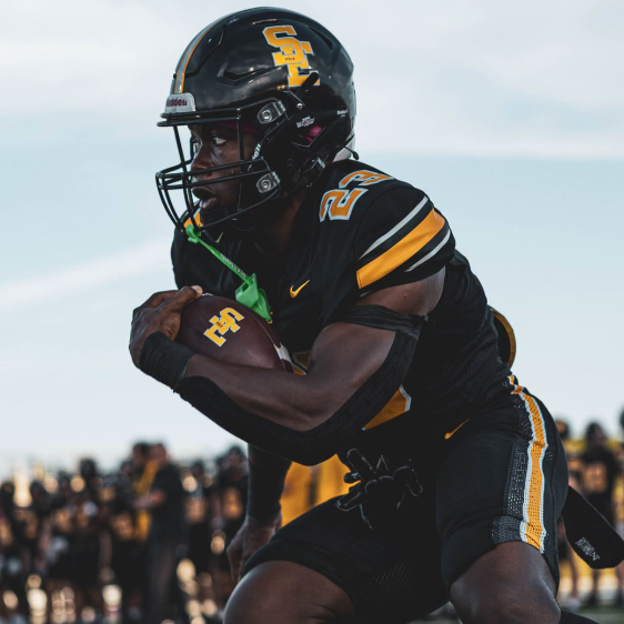 A football player in a black and yellow uniform wearing a helmet with a yellow “S” logo, arms crossed in front of the chest, standing on the field with a blurred crowd in the background.
