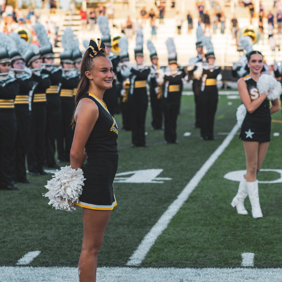 Cheerleaders in black and yellow uniforms perform on a football field, holding white pom-poms while marching and smiling as a line of band members in dark uniforms stands behind them.