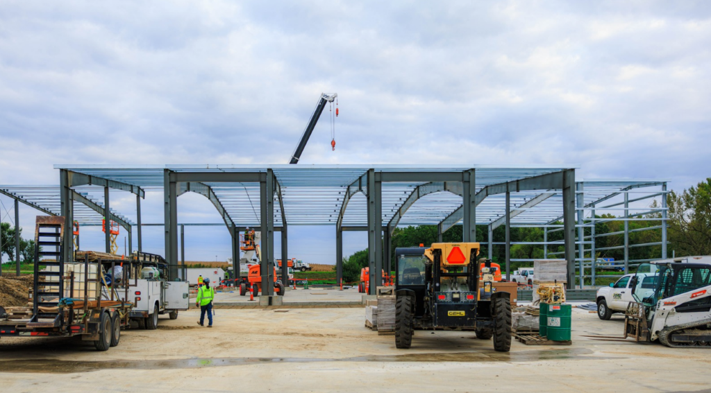 Construction site showing the structural steel framing of the new facility, with trucks, crew members, and an overhead crane against a cloudy sky.