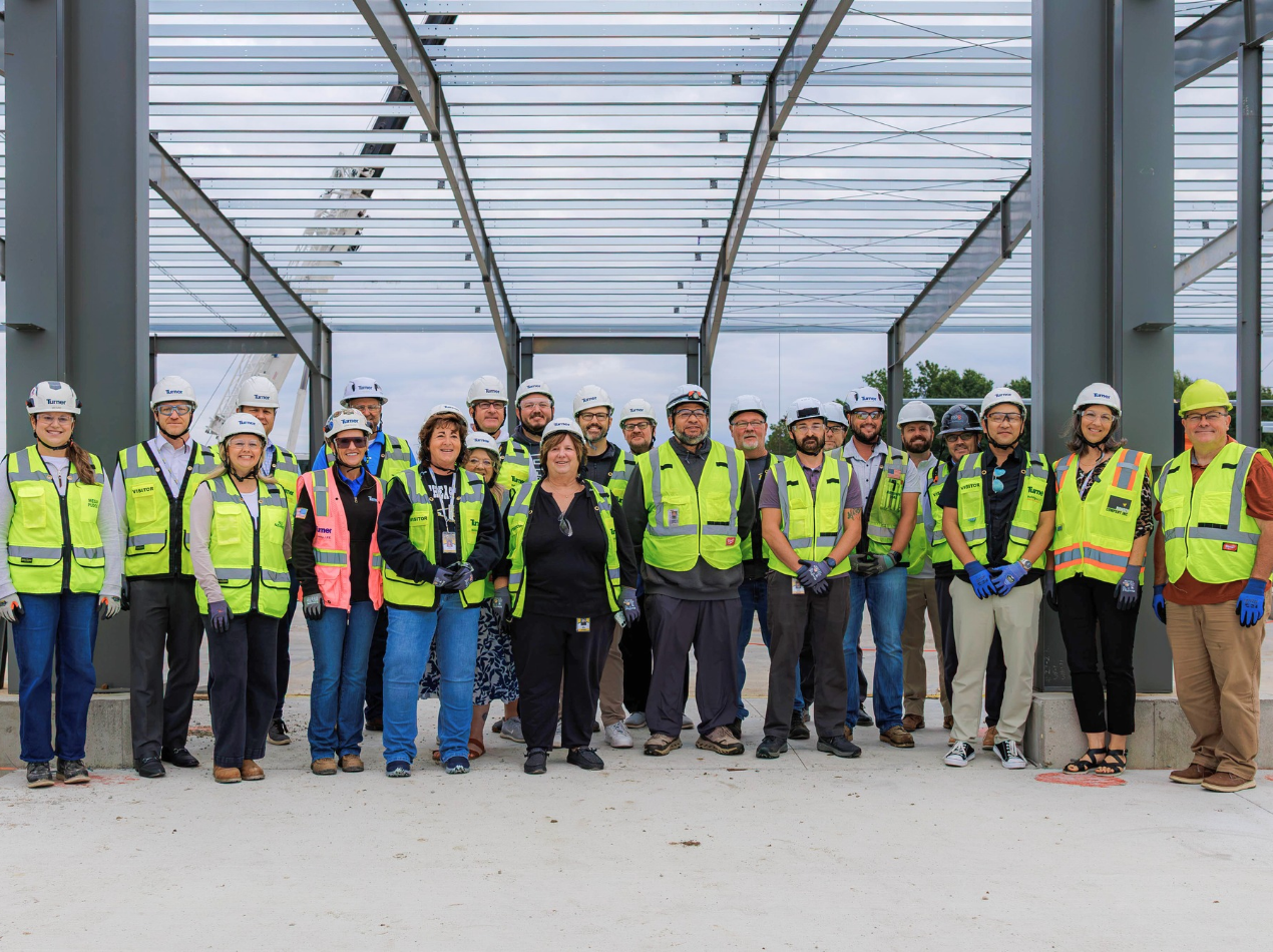 A group of construction workers, SEP staff, and engineers wearing hard hats and high-visibility vests stand in a line under a large steel structure with an open roof, posing for a group photo outdoors.